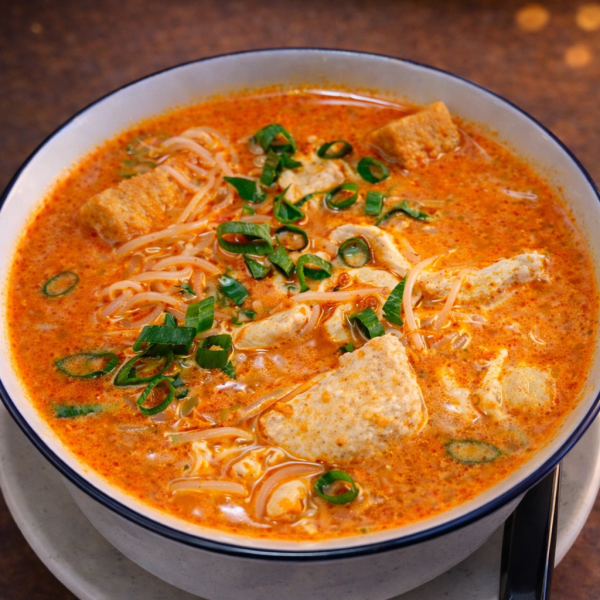 Bowl of spicy laksa noodle soup topped with chicken, tofu, and sliced scallions