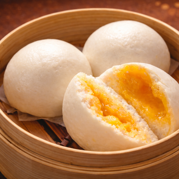 Three steamed buns in a bamboo steamer, with one bun broken open to reveal a molten salted egg yolk filling