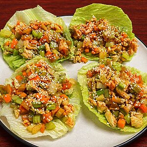 Four lettuce wraps filled with a stir-fry of diced chicken, carrots, and celery, topped with sesame seeds on a plate
