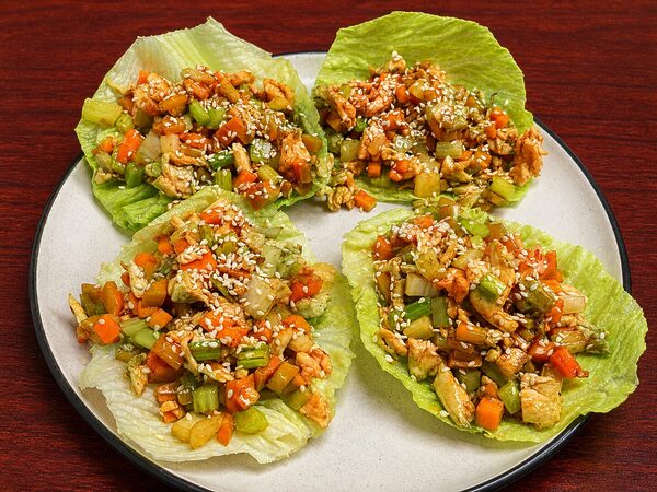 Four lettuce wraps filled with a stir-fry of diced chicken, carrots, and celery, topped with sesame seeds on a plate