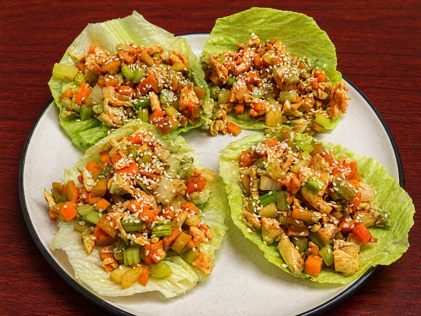 Four lettuce wraps filled with a stir-fry of diced chicken, carrots, and celery, topped with sesame seeds on a plate