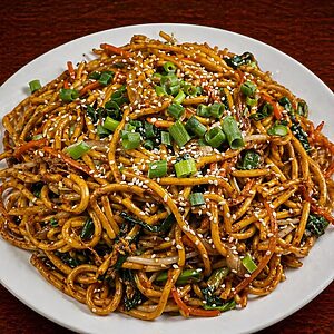 Stir-fried noodles with vegetables, garnished with sesame seeds and chopped green onions on a white plate