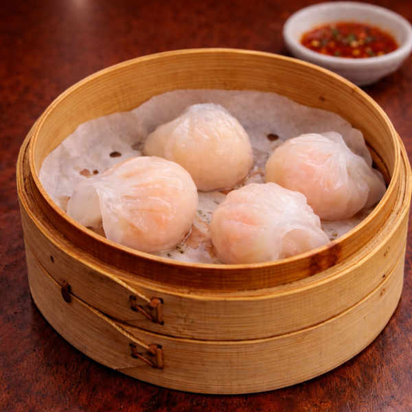 Four translucent shrimp dumplings served in a bamboo steamer with a side of chili sauce