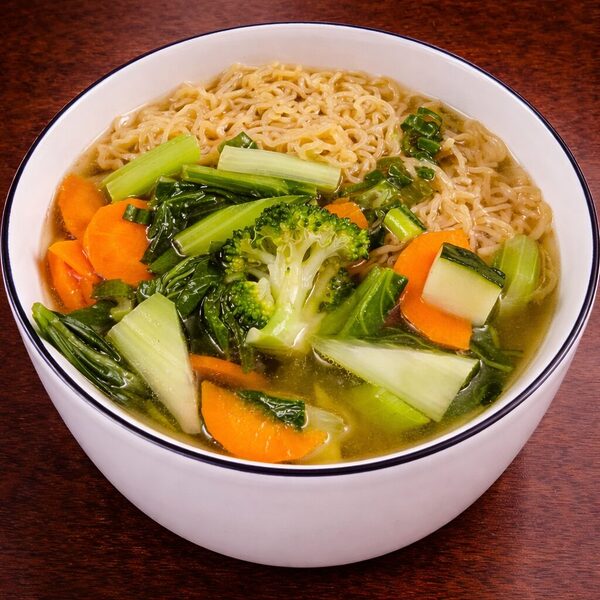 A bowl of vegetable noodle soup with broccoli, carrots, and leafy greens in a clear broth