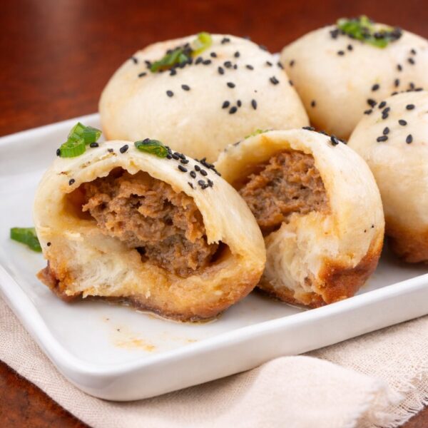 Pan-fried pork buns topped with black sesame seeds and green onions, with one bun split open to show the savory meat filling